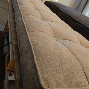 May include: A beige, tufted mattress with a rectangular shape. The mattress has a textured fabric and a stitched pattern creating a grid of squares. The edges are neatly finished with a contrasting trim. The mattress is placed on a wooden frame.