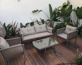 May include: Outdoor patio furniture set with a loveseat, two armchairs, and a coffee table. The furniture has a gray rope design with light beige cushions. The coffee table has a glass top. The set is on a wooden deck with plants in the background.