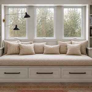 May include: A built-in window seat with a neutral color palette. The seat features a beige cushion and several decorative pillows in varying textures and patterns. Below the seat are three drawers with black handles. The window has a view of trees.
