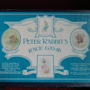Puede incluir: Una caja azul con un borde blanco y un panel central blanco. El panel central tiene el texto "Peter Rabbit's Race Game" en letras negras. La caja presenta ilustraciones de Peter Rabbit, Jemima Puddle-Duck, Squirrel Nutkin y Jeremy Fisher.