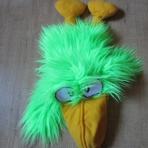 May include: A novelty bird costume head featuring bright green shaggy fur, a yellow beak, and two yellow head pieces. The eyes are white with black outlines. The item is on a wooden surface.