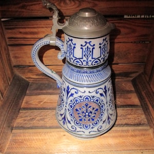 Puede incluir: Una jarra de cerveza de cerámica azul y blanca con una tapa de peltre. La jarra tiene un diseño floral y está decorada con detalles azules y dorados.