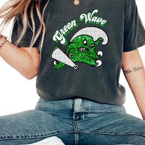 May include: A dark gray t-shirt with a green glitter graphic of a wave with a face and a megaphone. The text "Green Wave" is above the graphic.