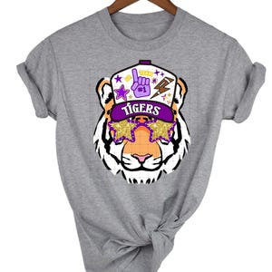 May include: Gray t-shirt with a cartoon tiger wearing a purple and gold baseball cap with the word "Tigers" on it. The tiger is wearing gold star-shaped sunglasses.