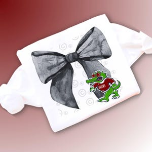 May include: White t-shirt featuring a large watercolor-style black bow and a cartoon alligator wearing a red shirt with the word "Gators". The alligator has green skin and is wearing star-shaped sunglasses.