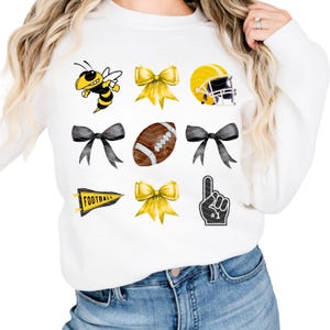 Puede incluir: Sudadera blanca con una variedad de gráficos deportivos amarillos y negros, incluyendo una abeja, lazos, un balón de fútbol, un casco de fútbol, un banderín que dice "FOOTBALL" y un puño en alto con el número "1" en él.