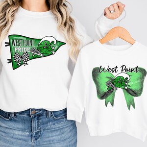 Puede incluir: Camisas blancas de manga larga con diseños gráficos verdes y negros. La camisa más grande tiene un banderín verde y negro con el texto "West Point Pride". La camisa más pequeña tiene un lazo verde y negro con el texto "West Point".