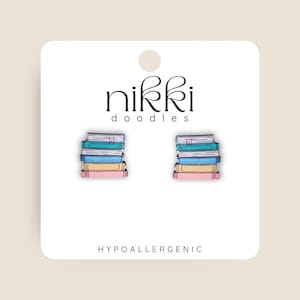 Peut inclure: Boucles d'oreilles hypoallergéniques représentant des piles de livres colorés. Chaque pile comprend des livres roses, jaunes, bleus et turquoise, avec un marque-page. La carte blanche porte le texte "nikki doodles".