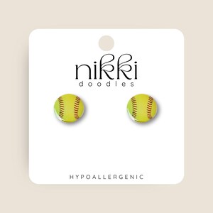 May include: A pair of hypoallergenic earrings featuring a yellow softball with red stitching. The earrings are packaged on a white card with the text "nikki doodles" and "HYPOALLERGENIC".