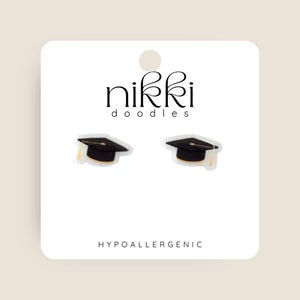 May include: A pair of hypoallergenic earrings featuring a black graduation cap design. The earrings are on a white card with the text "nikki doodles" and "HYPOALLERGENIC".