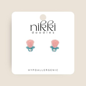 May include: A pair of pink and blue pacifier shaped earrings on a white card with the text "nikki doodles" and "HYPOALLERGENIC".