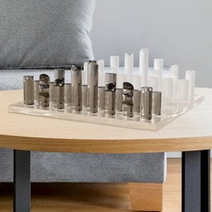 May include: A modern chess set with clear and gray pieces on a transparent board. The pieces are cylindrical and rectangular, arranged on a light wood table. The table has black legs and sits in a living room setting.