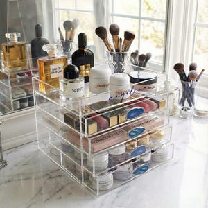 Puede incluir: Un organizador de maquillaje acrílico transparente con múltiples cajones y estantes, lleno de cosméticos, perfumes y brochas. El organizador está sobre una superficie de mármol, con un espejo en el fondo. Los cajones contienen barras de labios, paletas de sombras de ojos y productos para el cuidado de la piel.