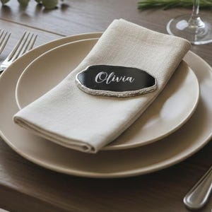 May include: Elegant table setting with beige plates, folded linen napkins, and silver cutlery. Each place setting includes a wine glass and a name card that reads "Olivia". The table is made of dark wood.