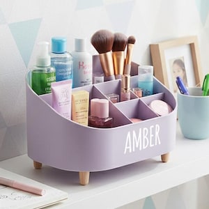 May include: A lavender cosmetic organizer with wooden legs, filled with makeup brushes, skincare products, and other beauty items. The name "AMBER" is printed in white on the front. The organizer sits on a white shelf with a notebook, pens, and a mug.