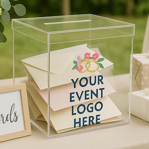 May include: A clear acrylic card box with a slot on top, filled with envelopes. The box has text that reads "YOUR EVENT LOGO HERE" and a floral design. A small framed sign that says "Cards" is next to the box. White flowers and gifts are in the background.