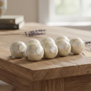 Könnte beinhalten: Acht dekorative Kugeln mit einem segmentierten, cremefarbenen und perlmuttartigen Design liegen auf einem hellbraunen Holztisch. Eine Vase und ein Lavendelzweig befinden sich im Hintergrund. Jede Kugel hat einen Durchmesser von ca. 4 cm.