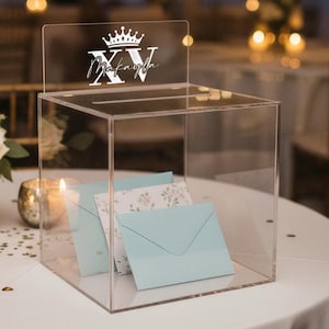 May include: Clear acrylic box with a slot on top, featuring a crown and the name "Makayla XV" in white script. Inside are light blue envelopes and floral-patterned cards. The box is on a white tablecloth.