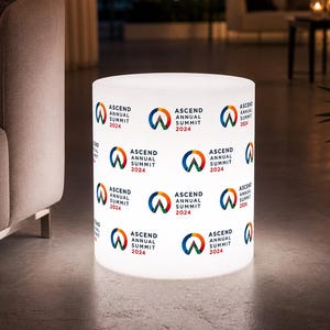 May include: A cylindrical, illuminated display stand with the text "ASCEND ANNUAL SUMMIT 2024" repeated around its surface. The logo features a stylized "W" in blue, orange, and green. The stand is white and appears to be indoors.