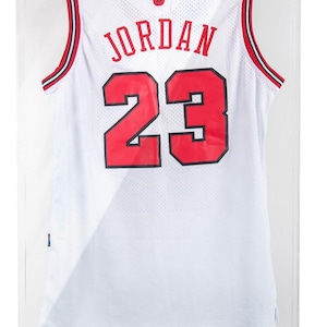 Puede incluir: Camiseta de baloncesto blanca con detalles rojos y negros, exhibida en una vitrina transparente. La camiseta muestra el nombre "JORDAN" y el n&uacute;mero "23" en rojo. El texto "MICHAEL JORDAN THE G.O.A.T." es visible en la parte inferior.