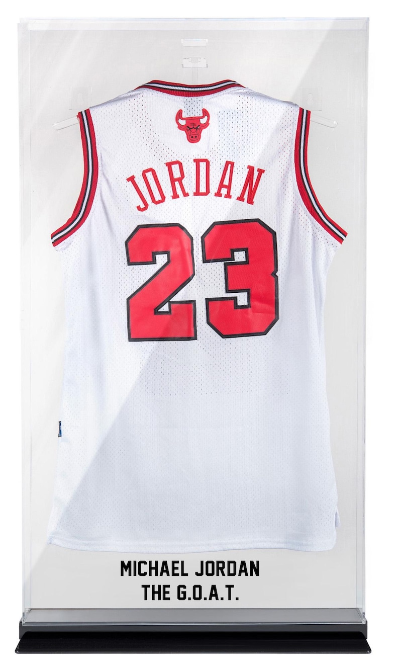 Puede incluir: Camiseta de baloncesto blanca con detalles rojos y negros, exhibida en una vitrina transparente. La camiseta muestra el nombre "JORDAN" y el n&uacute;mero "23" en rojo. El texto "MICHAEL JORDAN THE G.O.A.T." es visible en la parte inferior.