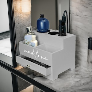 May include: A gray wooden organizer with multiple compartments and a drawer. The organizer holds bottles, a hair styling tool, and a comb. The name "Madeline" is written on the front in white script. The organizer is on a marble countertop.