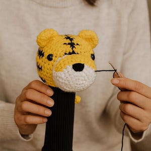 K&ouml;nnte beinhalten: Handgefertigte H&auml;kel-Tiger-Schl&auml;gerkopfhaube. Der Tiger hat einen gelben K&ouml;rper mit schwarzen Streifen, eine wei&szlig;e Schnauze und schwarze Augen. Die Schl&auml;gerhaube hat eine schwarze gerippte H&uuml;lle.