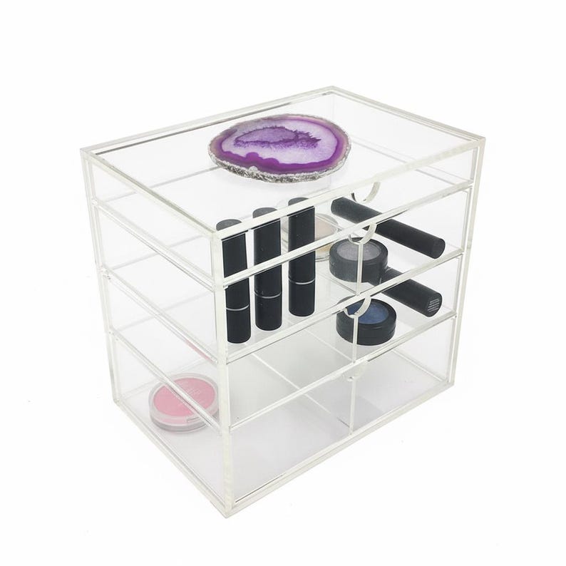 K&ouml;nnte beinhalten: Ein klarer Make-up-Organizer aus Acryl mit vier Schubladen. Auf der obersten Ablage befindet sich eine lila Geodenscheibe. Die Schubladen enthalten schwarze Lippenstifte, Lidschatten, Rouge und andere Kosmetikartikel.