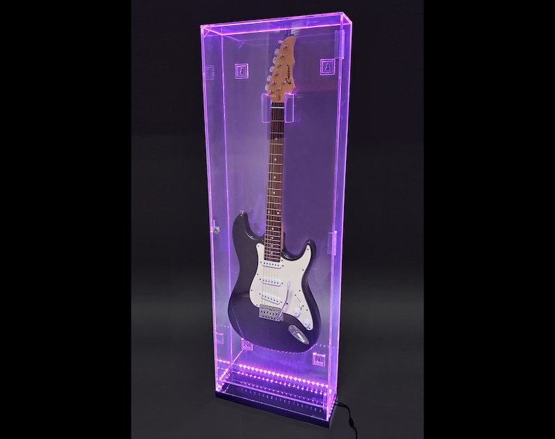 May include: A black electric guitar with a white pickguard is displayed inside a clear, rectangular case with purple LED lighting. The guitar case has a black base and a silver latch. The guitar's headstock is visible.