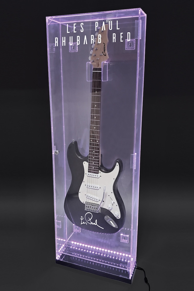 May include: A black and white electric guitar in a clear display case with the text "LES PAUL RHUBARB RED". The guitar has a signature on the body and is illuminated with purple lights.