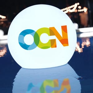 May include: A white illuminated sphere floating in a pool of water. The sphere has a colorful logo on it that says "OCN".