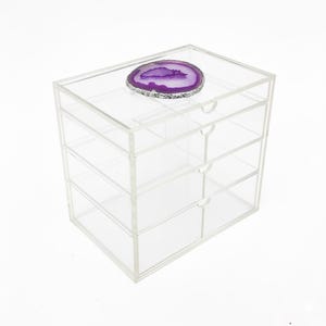 K&ouml;nnte beinhalten: Eine transparente Aufbewahrungsbox aus Acryl mit vier Schubladen. Auf der Oberseite liegt eine lila-silberne Geodenscheibe. Die Box hat kleine, runde Schubladengriffe. Das Gesamtdesign ist modern und minimalistisch.