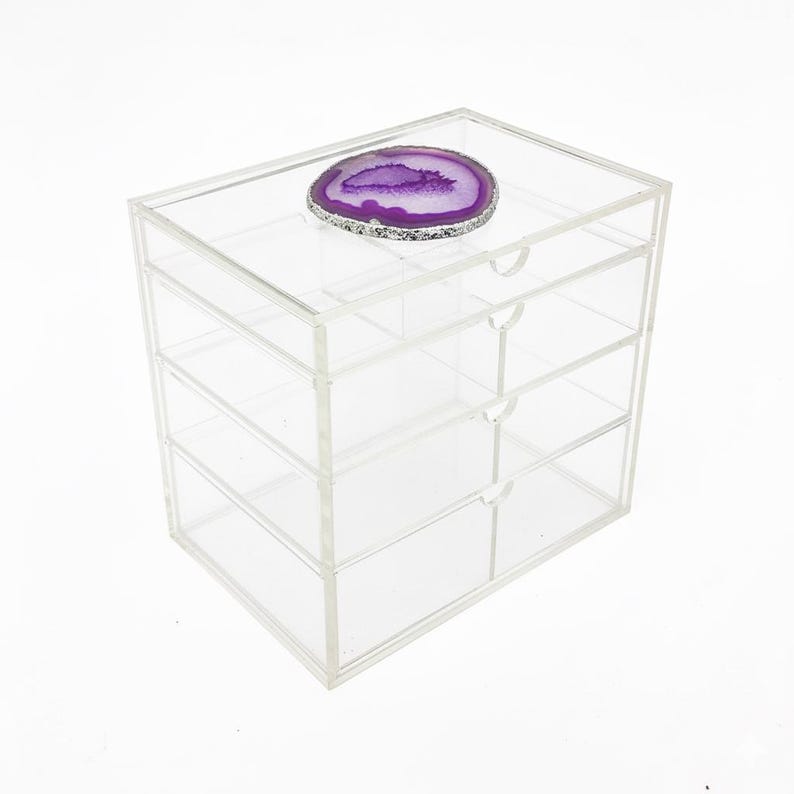 K&ouml;nnte beinhalten: Eine transparente Aufbewahrungsbox aus Acryl mit vier Schubladen. Auf der Oberseite liegt eine lila-silberne Geodenscheibe. Die Box hat kleine, runde Schubladengriffe. Das Gesamtdesign ist modern und minimalistisch.