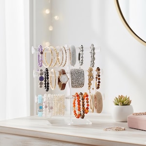May include: A clear acrylic bracelet display stand holding various bracelets. The bracelets are in a variety of colors, including gold, silver, orange, and purple. A brown leather watch is also displayed. The stand is on a light-colored wooden surface.