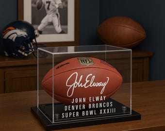 May include: A brown NFL football signed "John Elway" in a clear display case. The football is on a black base with text that reads "John Elway Denver Broncos Super Bowl XXXIII". A Denver Broncos helmet and a framed photo are in the background.