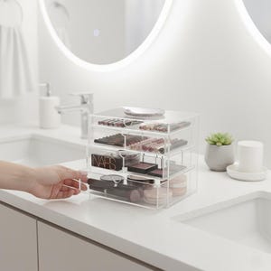 K&ouml;nnte beinhalten: Ein klarer Make-up-Organizer aus Acryl mit drei ausziehbaren Schubladen, gef&uuml;llt mit verschiedenen Kosmetika. Der Organizer steht auf einer wei&szlig;en Arbeitsplatte, wobei eine Hand eine der Schubladen herauszieht. Auf der Oberseite des Organizers befindet sich eine kleine, dekorative Schale.