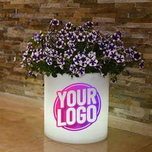 May include: White round planter with a purple and pink "YOUR LOGO" graphic, filled with purple and white flowers.