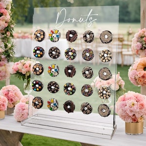 May include: A clear acrylic donut display stand with the word "Donuts" in script. The stand holds an assortment of decorated donuts with various toppings. Pink floral arrangements and a white table complete the scene.