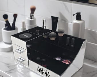 May include: A white and black makeup organizer with drawers and compartments, holding makeup brushes, compacts, and tubes. The organizer has the name "Corrine" in script on the side. The organizer is on a marble countertop.