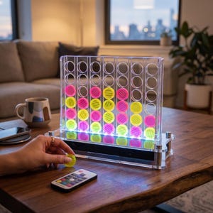 Op de afbeelding: Een vier-op-een-rij spelbord van helder acryl met verlichte sleuven die gele en roze ronde fiches bevatten. Het spel heeft een zwarte basis met geïntegreerde ledverlichting en wordt geleverd met een kleine afstandsbediening voor kleurinstellingen.
