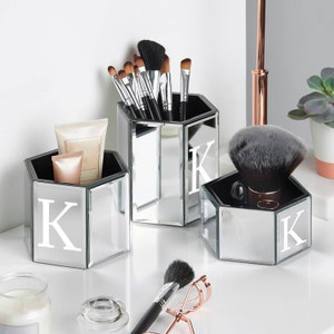 May include: Three mirrored hexagonal containers with black interiors and the letter "K" in white. The containers hold makeup brushes and beauty products. A rose gold eyelash curler and a makeup brush are in the foreground.