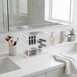 K&ouml;nnte beinhalten: Ein klarer Make-up-Organizer aus Acryl mit drei Schubladen, gef&uuml;llt mit Lippenstiften, Augenstiften und Make-up-Puder. Ein Keramikbecher enth&auml;lt Make-up-Pinsel. Eine kleine Sukkulentenpflanze steht daneben. Der Organizer steht auf einer wei&szlig;en Arbeitsplatte.