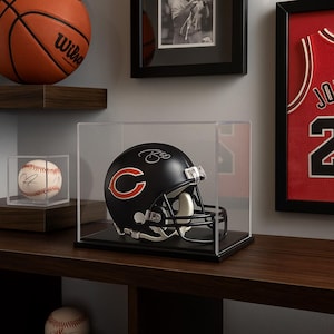 Könnte beinhalten: Ein schwarzer Chicago Bears Footballhelm mit orangefarbenem "C"-Logo, ausgestellt in einer transparenten Acrylvitrine. Der Helm steht auf einer schwarzen Basis und präsentiert Sportmemorabilien. Ein Basketball und ein Baseball sind ebenfalls sichtbar.