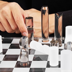 May include: A modern chess set with clear and metallic pieces on a black and white checkered board. The pieces have a minimalist design, with cylindrical and geometric shapes. The board is made of clear acrylic.