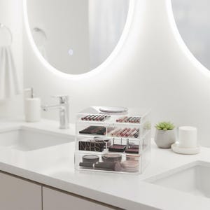K&ouml;nnte beinhalten: Ein klarer Make-up-Organizer aus Acryl mit mehreren Schubladen, gef&uuml;llt mit verschiedenen Kosmetika wie Lippenstiften, Lidschatten und Puder. Der Organizer steht auf einer wei&szlig;en Arbeitsplatte in einem Badezimmer.