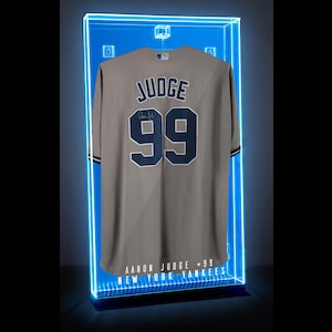 May include: A tan baseball jersey with the name "JUDGE" and the number "99" in navy blue. The jersey is displayed in a blue-lit display case. The text "AARON JUDGE - 99 NEW YORK YANKEES" is at the bottom.