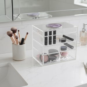 K&ouml;nnte beinhalten: Ein klarer Make-up-Organizer aus Acryl mit drei Schubladen, gef&uuml;llt mit Lippenstiften, Lidschatten und anderen Kosmetika. Der Organizer ist mit "amber" beschriftet. Make-up-Pinsel befinden sich in einem Keramikhalter.