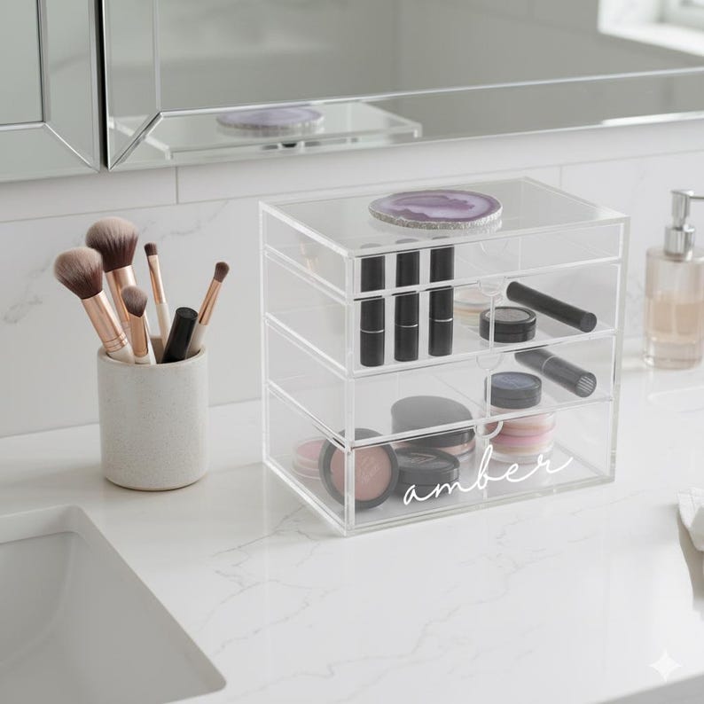 K&ouml;nnte beinhalten: Ein klarer Make-up-Organizer aus Acryl mit drei Schubladen, gef&uuml;llt mit Lippenstiften, Lidschatten und anderen Kosmetika. Der Organizer ist mit "amber" beschriftet. Make-up-Pinsel befinden sich in einem Keramikhalter.