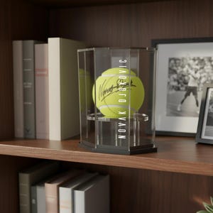 May include: A tennis ball in a clear display case. The ball is yellow with a signature and the name "NOVAK DJOKOVIC" printed vertically on the case. The case sits on a shelf with books and a framed picture.