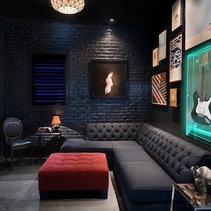 May include: A modern, dark-themed room with a black brick wall, a gray tufted sectional sofa, and a red ottoman. A guitar is displayed in a lighted acrylic case. Artwork and a small table with a lamp complete the space.